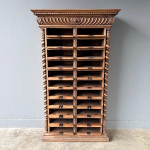 Puede incluir: Un armario alto de madera marr&oacute;n con dos columnas verticales de compartimentos de almacenamiento abiertos. La pieza est&aacute; decorada con detalles tallados acanalados en la cornisa superior y columnas de estilo soga trenzada a los lados.