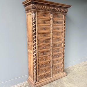 Puede incluir: Un armario de boticario alto de madera de color marr&oacute;n claro con dos columnas verticales de doce cajones cada una. La pieza est&aacute; adornada con intrincados detalles tallados, incluyendo columnas en espiral en los laterales y una cornisa decorativa.