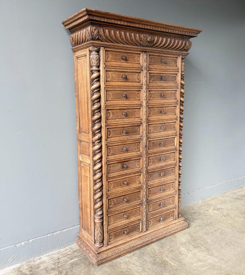 Puede incluir: Un armario de boticario alto de madera de color marr&oacute;n claro con dos columnas verticales de doce cajones cada una. La pieza est&aacute; adornada con intrincados detalles tallados, incluyendo columnas en espiral en los laterales y una cornisa decorativa.