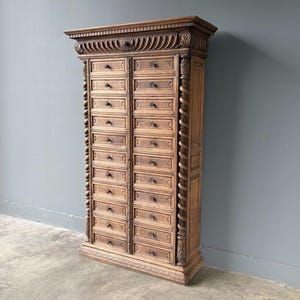 Puede incluir: Un armario de boticario alto de madera marr&oacute;n claro con dos columnas verticales de numerosos cajones peque&ntilde;os. Cada caj&oacute;n tiene un peque&ntilde;o pomo oscuro. El mueble est&aacute; decorado con columnas talladas en espiral y una moldura superior detallada.