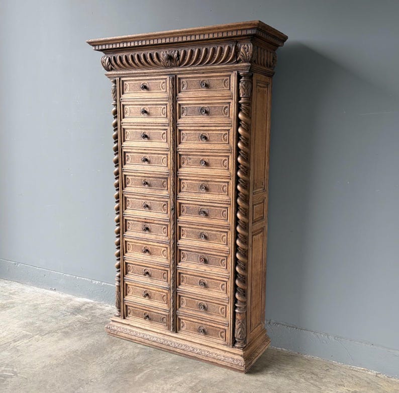 Puede incluir: Un armario de boticario alto de madera marr&oacute;n claro con dos columnas verticales de numerosos cajones peque&ntilde;os. Cada caj&oacute;n tiene un peque&ntilde;o pomo oscuro. El mueble est&aacute; decorado con columnas talladas en espiral y una moldura superior detallada.