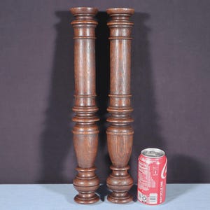 May include: Two antique, dark brown wooden columns with intricate carved details. Each column features a cylindrical body with decorative rings and a flared base. The columns are approximately 30 inches tall. A red soda can is next to the columns for size comparison.