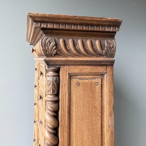 Puede incluir: Un armario alto de madera marr&oacute;n claro con detalles tallados intrincados, que incluyen una cornisa superior estriada, un friso decorativo con motivos de hojas y una columna vertical prominente con un dise&ntilde;o de espiral y follaje tallado.
