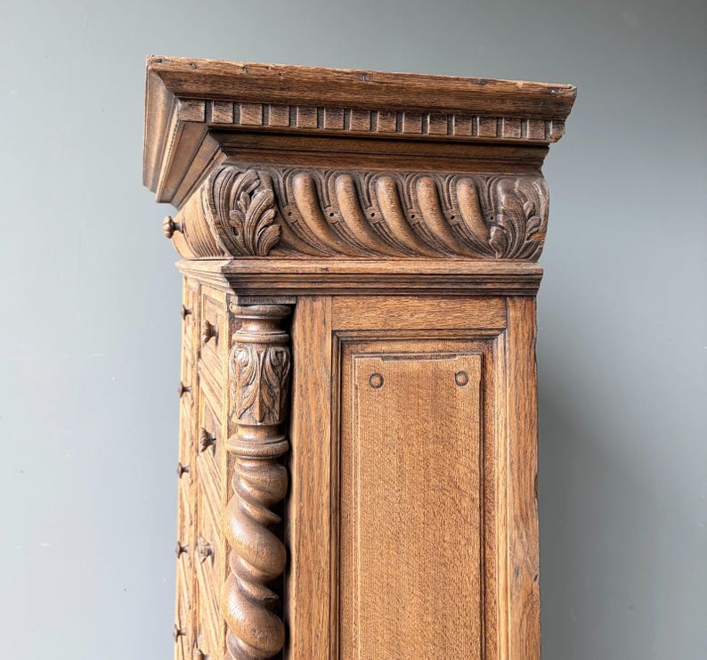 Puede incluir: Un armario alto de madera marr&oacute;n claro con detalles tallados intrincados, que incluyen una cornisa superior estriada, un friso decorativo con motivos de hojas y una columna vertical prominente con un dise&ntilde;o de espiral y follaje tallado.