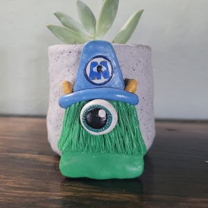 May include: A green and blue monster planter with a single eye and a blue hat with the letter "M" on it. The planter is made of ceramic and has a textured surface.