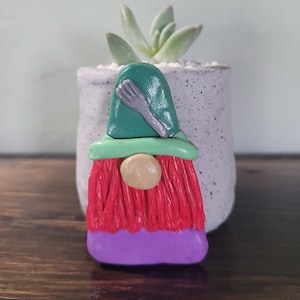 May include: A colorful gnome figurine with a green hat and a red beard. The gnome has a tan nose and a purple base. A silver fork and knife are on the hat. The gnome is in front of a gray pot with a succulent plant.