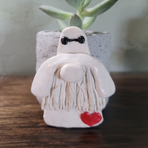 May include: White ceramic gnome with a red heart, wearing a white beard and black eyes. The gnome is sitting in a gray concrete planter with a green succulent plant.
