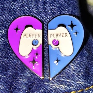 May include: A heart-shaped enamel pin with a purple and blue design. The pin features two video game controllers with the text "Player 1" and "Player 2" on each side.
