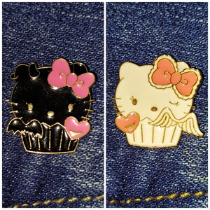 Può includere: Due spille a forma di cupcake a tema Hello Kitty. Una è nera con ali di pipistrello e un fiocco rosa. L'altra è bianca con ali d'angelo e un fiocco rosa.