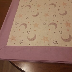 May include: A white fabric baby blanket with a pink border. The blanket features a repeating pattern of stars and moons.
