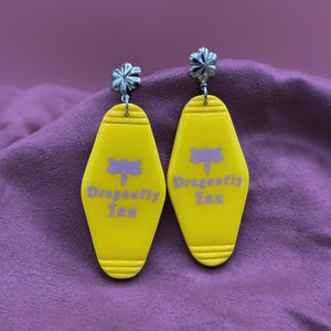 May include: A pair of yellow acrylic earrings shaped like motel keychains. Each earring features a purple dragonfly graphic and the text "Dragonfly Inn". The earrings have silver-colored floral studs.