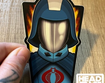Könnte beinhalten: Ein blauer und silberner Aufkleber mit einer Figur aus der G.I. Joe-Zeichentrickserie. Die Figur trägt einen blauen Helm und einen blauen Umhang mit einem Cobra-Symbol auf der Brust. Der Aufkleber hat einen weißen Hintergrund mit einem gelben und orangefarbenen Flammenmuster hinter der Figur. Der Aufkleber trägt den Text "HEADSPACE GRAPHICS" in der rechten unteren Ecke.