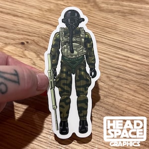 May include: A green and brown camouflage military action figure with a black helmet and gas mask. The figure is holding a green rifle and is standing on a white background.