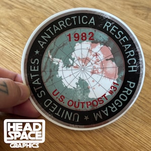 May include: A round sticker with a map of Antarctica in green and red, surrounded by text that reads "STATES ANTARCTICA RESEARCH PROGRAM" and "U.S. OUTPOST #31 1982." The sticker has a white border and the Head Space Graphics logo.
