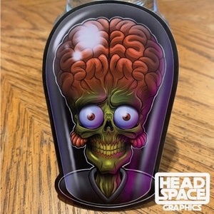 May include: A sticker featuring a cartoon illustration of a green alien with a large brain, big eyes, and a toothy grin. The alien is inside a clear dome, and the sticker has a black background with the text "HEADSPACE GRAPHICS" in white.