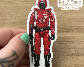 Könnte beinhalten: Eine rote und schwarze Actionfigur eines Soldaten mit einer Waffe. Die Figur trägt einen Helm und hat ein Cobra-Logo auf der Brust. Die Figur steht auf einem weißen Hintergrund.