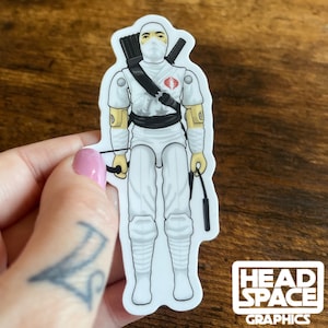May include: White vinyl sticker of a cartoon action figure wearing a white ninja suit with a black belt, a red Cobra symbol on his chest, and a yellow headband. He is holding a black whip in his right hand and a bow and arrow in his left hand. The sticker is on a wooden surface.
