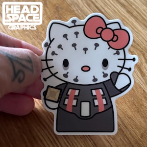 May include: A white Hello Kitty sticker with a black and red Darth Vader costume, a red bow, and black pins on her head.
