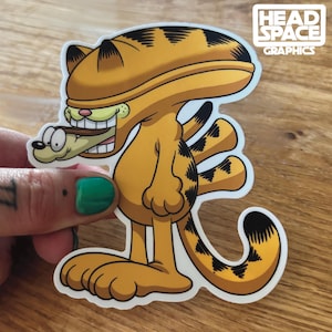 May include: A die-cut sticker featuring a cartoon Garfield character in a costume, with a dog in its mouth. The sticker is orange with black stripes and accents. The text "HEAD SPACE GRAPHICS" is in the upper right corner.
