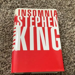 May include: A hardcover book titled "Insomnia" by Stephen King. The book has a red cover with the title and author's name in large white letters. The book is closed and lying flat.