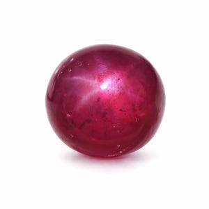 May include: A round, polished ruby gemstone with a star effect. The gem is a deep, saturated red color with a subtle, six-rayed star pattern visible on its surface. The ruby is set against a white background.