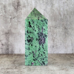 Op de afbeelding: Een groene en zwarte zoisiet kristallen toren met een driehoekige top. De steen heeft een gespikkeld patroon met donkergroene en zwarte vlekken. Het kristal staat rechtop op een grijs oppervlak.