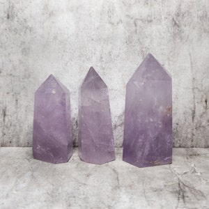 Op de afbeelding: Drie amethistkristal obelisken van verschillende groottes. De kristallen zijn een doorschijnende paarse kleur met zichtbare interne structuren. Elke obelisk heeft een puntige top en een rechthoekige basis. De achtergrond is een gevlekt grijs.