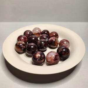 Op de afbeelding: Een verzameling gepolijste, ronde edelstenen met een mix van rode, bruine en heldere insluitsels, gepresenteerd in een crèmekleurige, ondiepe kom. De edelstenen hebben een reflecterend, glanzend oppervlak.