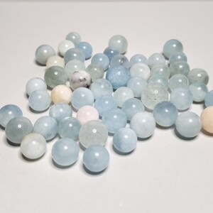 Op de afbeelding: Een verzameling aquamarijn edelsteen kralen in verschillende tinten blauw, groen en wit. De bolvormige kralen zijn verspreid en tonen hun gepolijste oppervlakken en natuurlijke variaties in kleur en textuur. Ideaal voor het maken van sieraden.