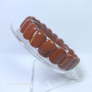 Può includere: Un braccialetto in diaspro rosso con perline rettangolari. Le perline presentano variazioni naturali di colore e motivo, alcune con striature marrone chiaro e crema. Il braccialetto è esposto su un supporto trasparente e curvo.