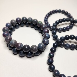 Op de afbeelding: Verschillende armbanden met kralen van donkerblauwe, grijze en kastanjebruine kralen. De armbanden zijn gerangschikt op een wit oppervlak, met kralen van ongeveer 1 cm in diameter. De armbanden zijn waarschijnlijk gemaakt van natuursteen.