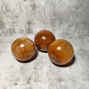 Op de afbeelding: Drie gepolijste, bolvormige edelstenen in tinten amber en bruin. De bollen hebben een glad, reflecterend oppervlak, met zichtbare interne patronen. De stenen zijn gerangschikt op een gestructureerd, grijs oppervlak.