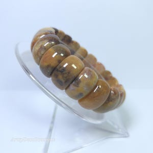 Op de afbeelding: Een rekbare armband met grote, afgeronde kralen in bruin-, beige- en crèmetinten. De kralen hebben een gemarmerd uiterlijk en zijn aan elkaar geregen. De armband wordt gepresenteerd op een heldere, schuine standaard.