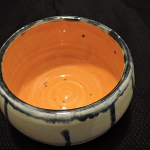 Könnte beinhalten: Eine kleine Keramikschüssel mit weißem Äußeren und schwarzen Glasur-Akzenten. Das Innere der Schüssel ist leuchtend orange.