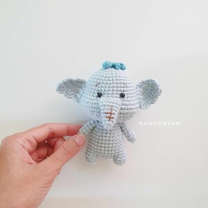 May include: A light blue crocheted elephant toy with black eyes and a brown nose. The elephant has a blue crocheted bow on its head. NAMSOMPAN is written on the white background.