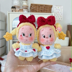 Könnte beinhalten: Zwei handgefertigte Häkelpuppen mit rosa Gesichtern, gelbem Haar und großen roten Schleifen. Jede Puppe trägt ein hellblau-weiß kariertes Kleid mit kleinen roten Herzakzenten und gelben sternförmigen Details. Der Text @NAMSOMPAN ist sichtbar.