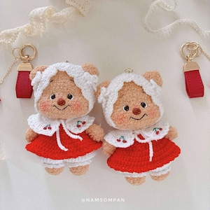 May include: Two handmade crochet bear keychains. Each bear is dressed in a red dress with a white collar and a matching bonnet. The keychains have gold-colored hardware and red leather wrist straps. The bears have embroidered facial features.