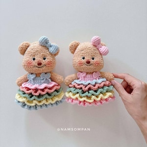 以下が含まれることがあります： 手作りのかぎ針編みテディベア2体。各クマは、薄茶色の体、頭の上のリボン、パステルカラーのフリルドレスを着ています。クマには刺繍された顔の特徴とバラ色の頬があります。