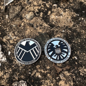 Peut inclure: Deux pièces rondes noires et argentées avec l'inscription "STRATEGIC HOMELAND INTERVENTION ENFORCEMENT LOGISTICS DIVISION". L'une représente un bouclier, l'autre un aigle.