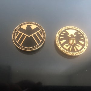 May include: Two gold-coloured metal coins with black and gold designs. One coin features a stylized eagle and shield, the other a geometric emblem. Both coins have the text "STRATEGIC HOMELAND INTERVENTION ENFORCEMENT LOGISTICS DIVISION".