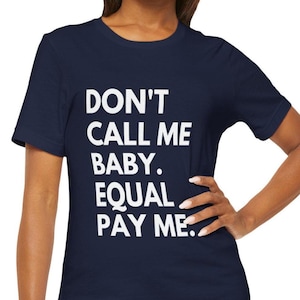 May include: Navy blue t-shirt with the text "DON'T CALL ME BABY. EQUAL PAY ME." in white block letters. The shirt is a short-sleeved crew neck style.