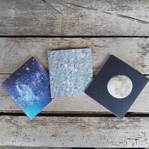 May include: Three square cards with different space-themed designs. The first card has a blue and purple nebula with stars. The second card has a gray and white image of the moon's surface. The third card has a dark blue background with a white moon.