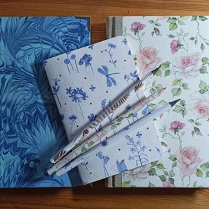 May include: Four notebooks with different patterned covers, including blue marbled, floral, and blue dragonfly designs. Three pencils are placed on top of the notebooks.
