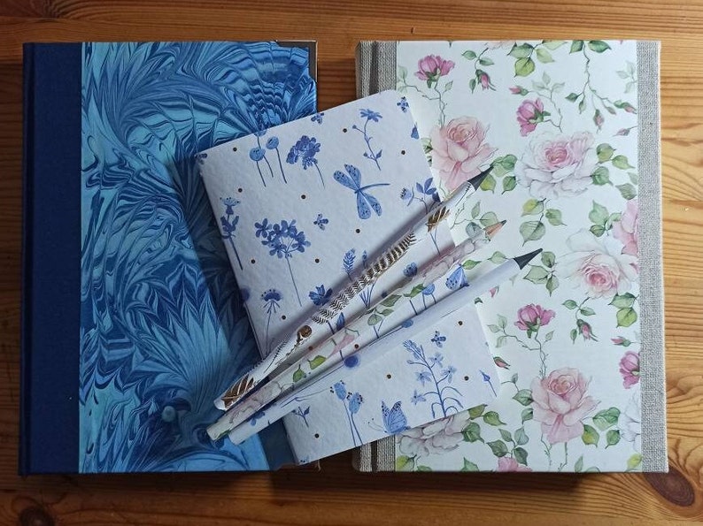 May include: Four notebooks with different patterned covers, including blue marbled, floral, and blue dragonfly designs. Three pencils are placed on top of the notebooks.