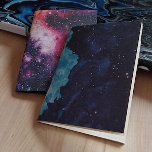May include: Two notebooks with galaxy-themed covers. One cover features a pink and purple galaxy with white stars, the other a dark blue galaxy with white stars and a teal watercolor effect.
