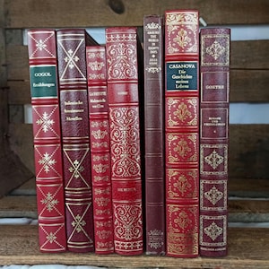 May include: A collection of eight vintage hardcover books with red leather covers and gold lettering. The titles include "Gogol", "La Frette", "Corry", "The World in Eighty Days", "Casanova", "Die Geschichte meines Lebens", and "Goethe".