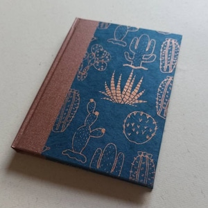 Puede incluir: Un cuaderno azul verdoso con una encuadernación de color cobre. La portada presenta un patrón repetido de cactus y suculentas en una impresión de lámina de cobre.
