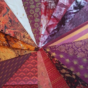 May include: A collection of patterned papers in shades of red, orange, and purple. The papers are arranged in a radial pattern, with the center of the pattern being at the top of the image. The papers have a variety of patterns, including marbled, geometric, and floral.