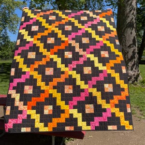Könnte beinhalten: Ein farbenfroher Quilt mit einem geometrischen Muster. Der Quilt hat einen schwarzen Hintergrund mit einem sich wiederholenden Design aus orangefarbenen, gelben und rosa Blöcken. Der Quilt wird im Freien auf einem roten Picknicktisch ausgestellt.
