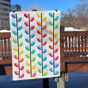 May include: A colorful quilt with a white background and a repeating pattern of rainbow-colored leaf shapes. The leaves are arranged in vertical rows with black lines. The quilt is hanging outdoors on a wooden structure.
